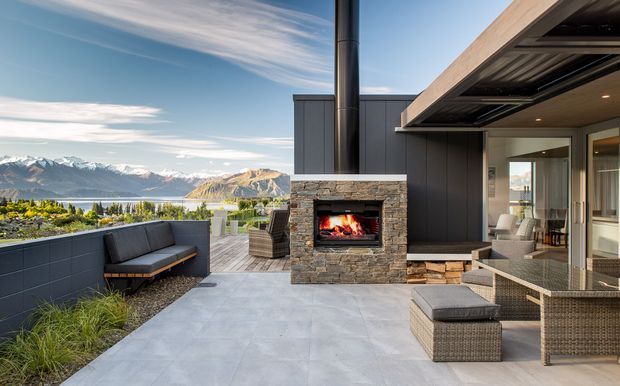 A contemporary outdoor terrace overlooking a lake and dramatic mountain landscape. The space features a built-in stone-clad fireplace with a live fire, dark vertical-clad walls, broad concrete paving, a covered roof extension, and woven outdoor lounge furniture with an integrated bench and planters.