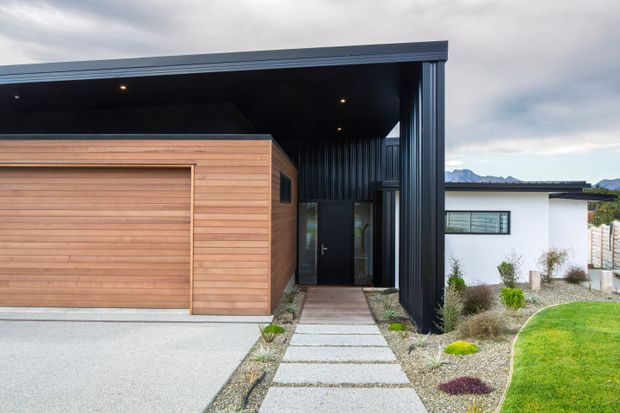 Contemporary residential exterior featuring a dark metal-clad flat-roofed entry structure, warm horizontal timber-clad garage volume, concrete stepping-stone pathway, and minimalist landscaped planting. A white single-storey wing and distant mountain backdrop are visible beyond.