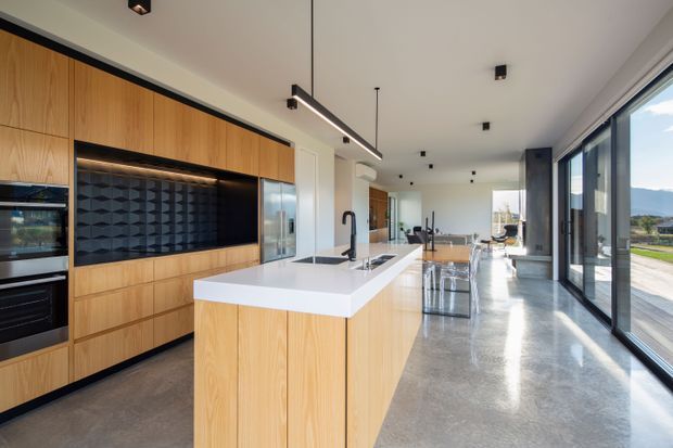 A spacious contemporary kitchen with a long central island, white stone benchtop, timber veneer cabinetry, integrated black appliances, and a dark geometric feature splashback. Full-height glazing along one side brings in abundant natural light and opens to a landscaped outdoor setting. Polished concrete flooring and linear pendant lighting contribute to the refined modern aesthetic.