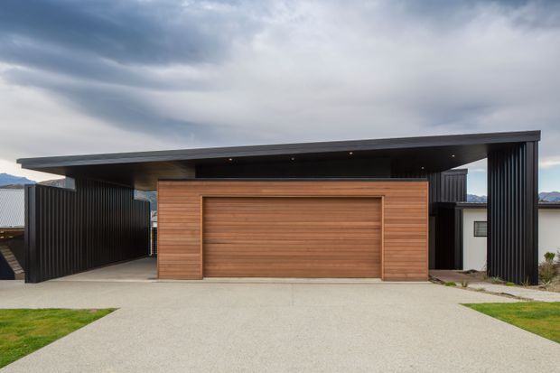 Front exterior view of a contemporary single-storey residence with a broad flat roof and deep cantilevered carport. The facade combines dark vertical metal screening with warm horizontal timber garage cladding, opening onto a wide concrete driveway.