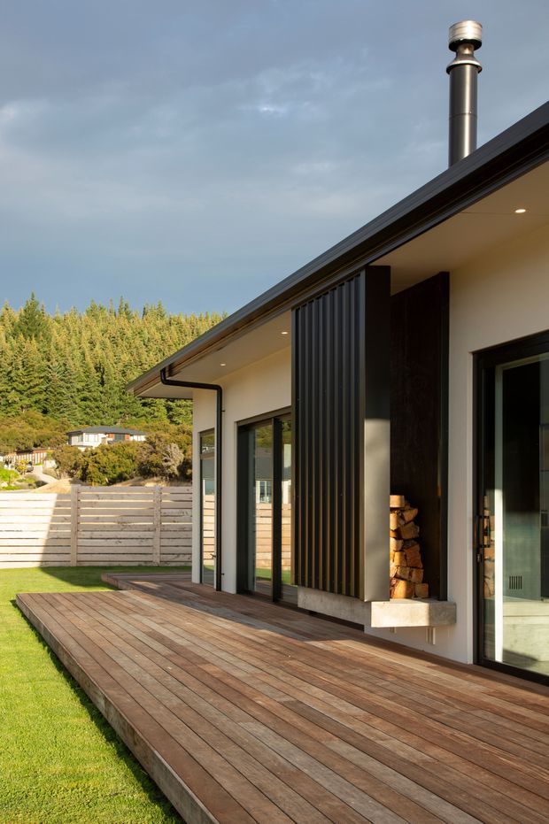 Exterior view of a contemporary single-storey home with a broad timber deck, dark metal roofing and window frames, pale rendered walls, vertical dark cladding, and a prominent metal chimney. Large glazed doors open toward the deck, with a built-in outdoor fireplace or wood storage niche. The house sits in a landscaped suburban setting backed by dense trees and neighbouring homes.