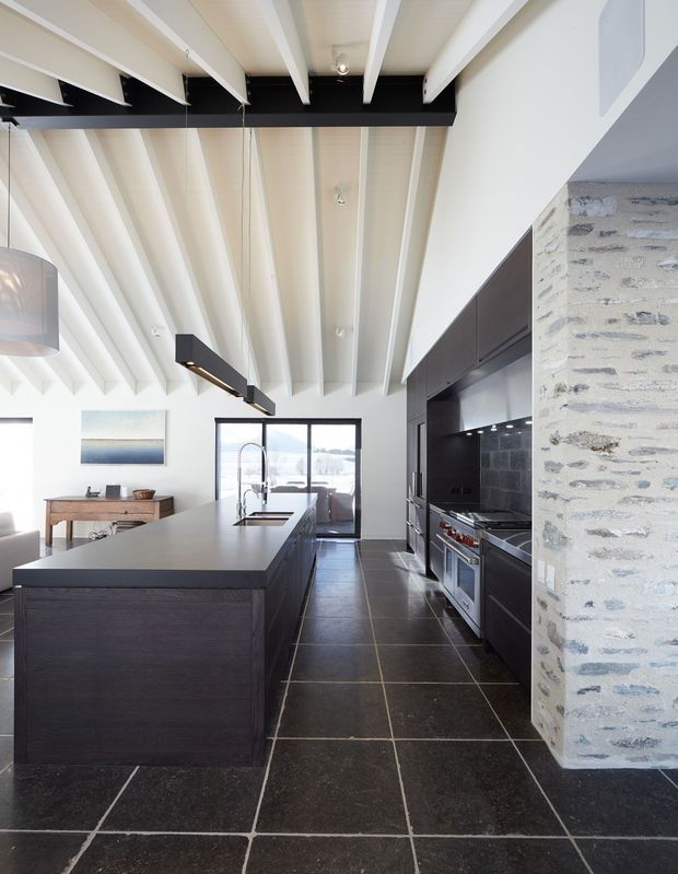 Bright contemporary kitchen with a large dark timber island, integrated sink and tap, dark cabinetry, stainless-steel appliances, black tiled backsplash, and large dark floor tiles. Exposed white ceiling beams create a strong architectural feature, while a rough pale brick column separates the kitchen from the adjacent dining/living area. Glazing at the rear opens toward an outdoor view.