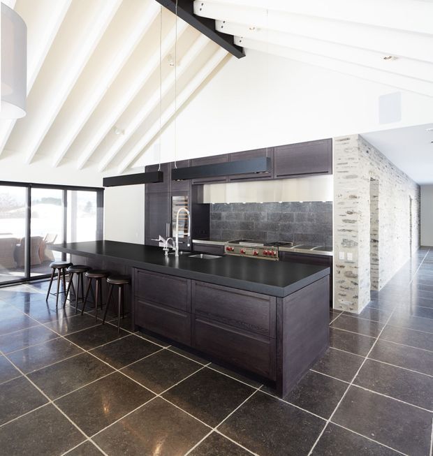 A spacious contemporary kitchen with a large dark timber island, black benchtop, integrated cooktop, sink and breakfast bar seating. A dark stone or tiled splashback sits beneath streamlined upper cabinetry, while white exposed roof beams and expansive glazing create a bright, open interior. The floor is finished in large dark square tiles, and a pale textured masonry wall defines the adjacent space.