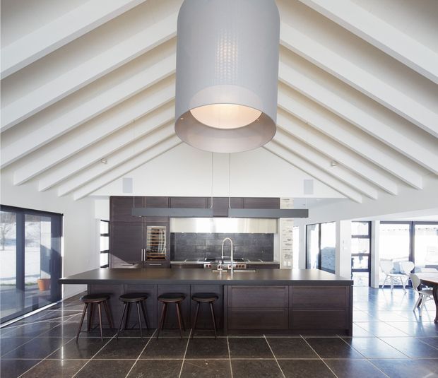 A bright, open-plan kitchen dominated by a large dark island with integrated cabinetry and seating for four. The kitchen features a dark stone-look splashback and benchtop, stainless-steel appliances and tapware, extensive glazing, and a dramatic white vaulted ceiling with exposed rafters. A large cylindrical pendant light hangs centrally above the island, while a dining or living area is visible beyond the kitchen.