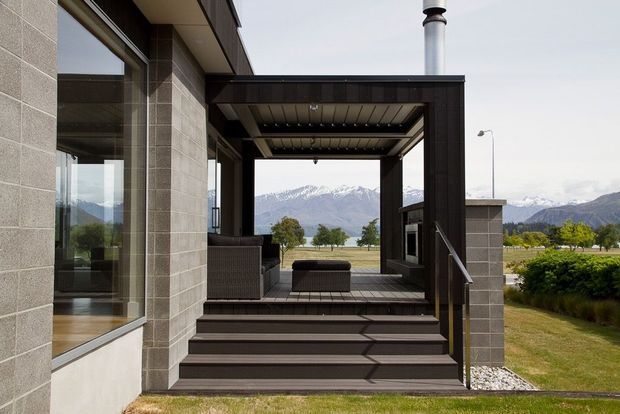 Contemporary covered outdoor terrace or entry deck attached to a modern home, with broad steps, dark timber decking and framing, a roofed outdoor room, built-in seating, and a visible chimney. Large glazing and pale masonry walls connect the terrace to the interior. The setting opens toward a rural landscape and snow-capped mountains.