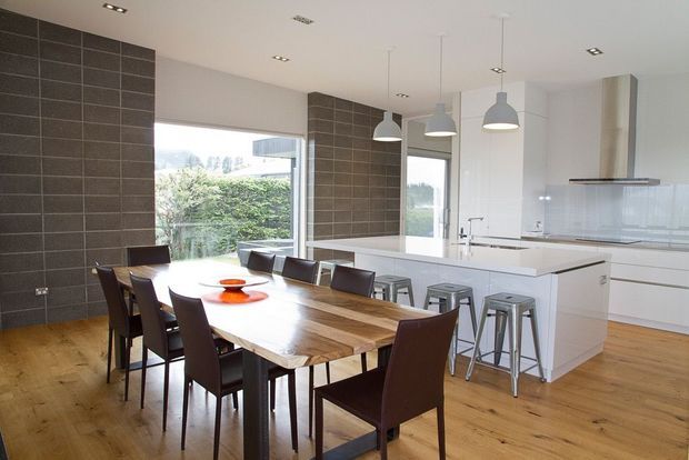 Bright contemporary open-plan kitchen and dining area with a long solid timber dining table, dark upholstered dining chairs, a white island with metal bar stools, white handleless cabinetry, dark tiled feature walls, timber flooring, pendant lights, and large windows overlooking greenery.