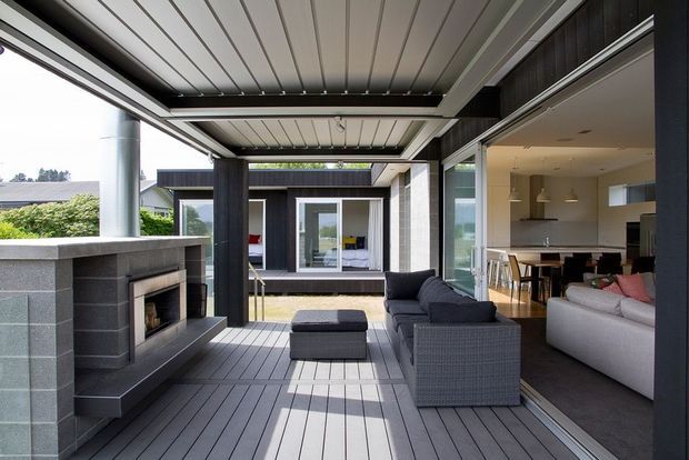 Contemporary covered outdoor living area with a grey composite or timber-look deck, built-in masonry fireplace, dark structural framing, and lounge seating. Large sliding glass openings connect the terrace directly to a bright open-plan living and kitchen area, with a landscaped suburban setting visible beyond.