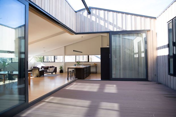 A bright contemporary courtyard view of a modern home, with a large timber deck in the foreground and an open-plan kitchen and living area beyond. The building has extensive floor-to-ceiling glazing, dark aluminium-framed sliding doors, and pale vertical weatherboard cladding. The interior includes a central kitchen island, timber flooring, dining and lounge furniture, and high clerestory-style windows.