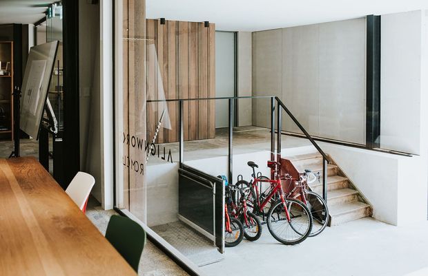 A contemporary interior showing a timber dining table and chairs in the foreground, opening onto a split-level circulation area with pale concrete or stone steps, black metal and glass balustrades, timber wall panelling, and bicycles stored below the stairs. The space has a clean, open, utilitarian character with strong natural materials and glazing.