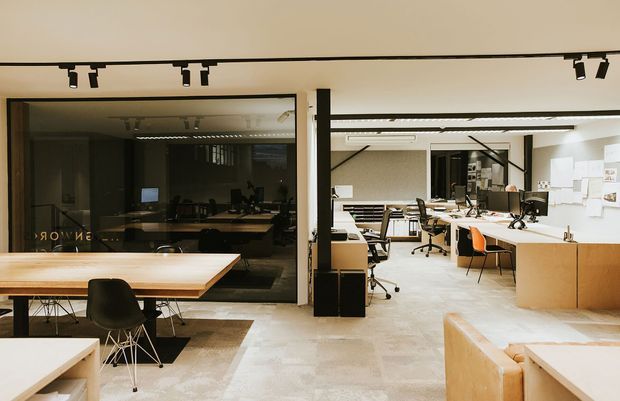 A bright contemporary open-plan design studio or office with multiple workstations, large desks, task chairs, wall-mounted drawings and shelving. The space has polished concrete flooring, light walls, timber-toned cabinetry and counters, black-framed glazing, and exposed track lighting. A dark meeting room is visible behind the internal glass partition, while a lounge or seating area occupies the foreground.