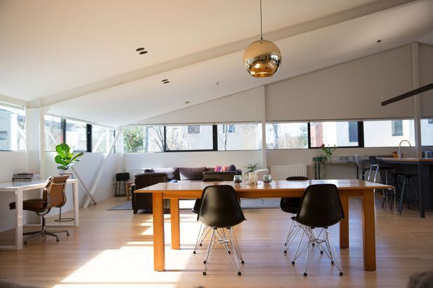 Bright open-plan interior with a large timber dining table and modern dining chairs in the foreground, a lounge area beyond, and a compact kitchen with island seating at the rear. Full-width horizontal windows, pale timber flooring, white walls and ceiling, and a reflective spherical pendant create an airy contemporary living space.
