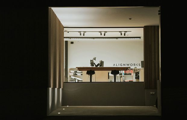 A dark, dramatically framed view through a large interior opening into a contemporary design studio or commercial workspace. Vertical timber slats line both sides of the opening, with a deep grey lower wall, recessed floor lights, a long timber work counter, stools, shelving, and track lighting. The word “ALIGNWORKS” is visible on the rear wall.
