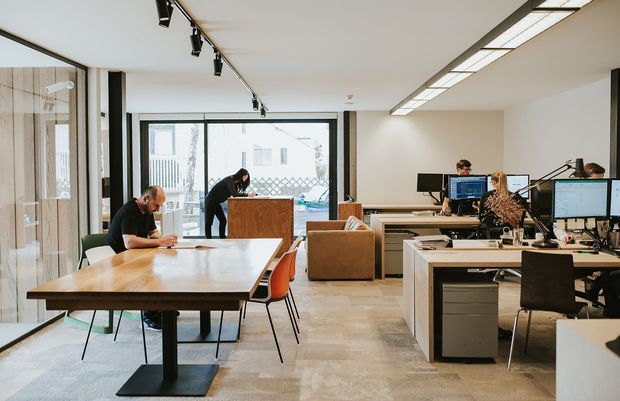 A bright, open-plan contemporary office interior with multiple workstations, desktop computers, a large communal timber worktable, and a timber reception or kitchenette counter. The space has polished concrete flooring, extensive glazing, exposed black track lighting, linear ceiling lights, and a small lounge area with a tan sofa. Several people are working at desks.