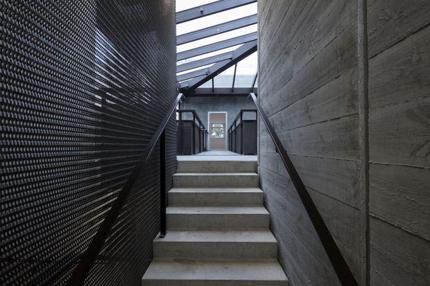 A narrow, dramatic interior stairway rises between a rough board-formed concrete wall and a dark perforated metal screen wall. Raw concrete steps, slim dark handrails, black metal balustrades, and a glazed overhead roof create an industrial, linear passage leading to a doorway.