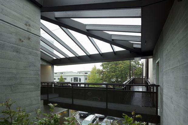 A covered elevated outdoor walkway or terrace framed by exposed dark steel beams and translucent sloped glazing. Rough horizontal concrete walls, black metal balustrades, and planted edges create a robust contemporary architectural setting, with an urban building and parked cars visible beyond.