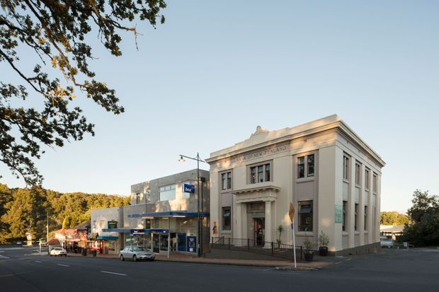 Street-level exterior view of a formal, classical-style civic or commercial building on a suburban town-centre intersection. The prominent cream-painted masonry facade has a symmetrical composition, pilasters, tall sash-like windows, a prominent cornice and classical entrance surround. Adjacent low-rise commercial buildings and streetscape elements are visible.