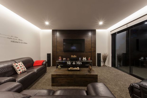 A contemporary media room with dark leather sofas and armchairs arranged around a low timber coffee table and a wall-mounted television. The TV wall features dark wood-look paneling, built-in display shelving, and decorative objects, while recessed and cove lighting create a warm atmosphere. Floor-to-ceiling dark-framed glazing occupies the right side, and a carpeted floor spans the room.