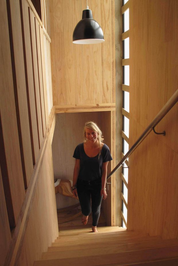 A narrow, warmly lit interior stairway lined with natural timber wall panelling and timber treads. A black pendant light hangs above the stairwell, while a tall vertical strip window with timber framing brings in daylight. A simple dark handrail runs along the right wall, and a person is walking up the stairs.