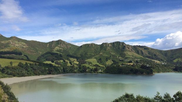 A scenic landscape view of a calm lake or reservoir surrounded by steep, green, forested mountains and rolling rural hills. The image is dominated by natural terrain, water, and sky, with sparse distant settlement visible along the hillsides.