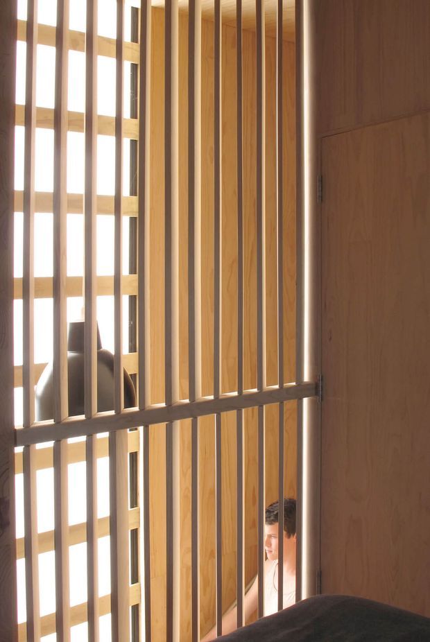 Interior architectural photograph dominated by a warm timber slatted screen or balustrade, with vertical wood battens, horizontal supports, and a timber-paneled door or wall at right. Bright glazing behind the screen creates strong filtered daylight. Partial views of a seated person and dark upholstered furniture are visible beyond the screen.