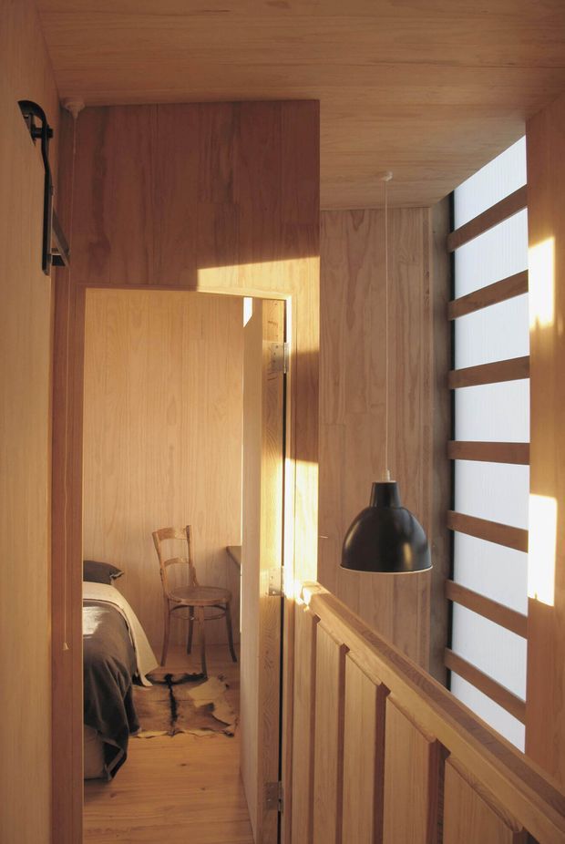 A warm, compact timber-lined landing or hallway viewed beside an open bedroom door. The space features extensive natural wood wall and ceiling panelling, timber balustrades, a black pendant light, and a tall window with horizontal timber battens filtering bright sunlight. A bedroom with a bed, chair, and small rug is visible through the doorway.