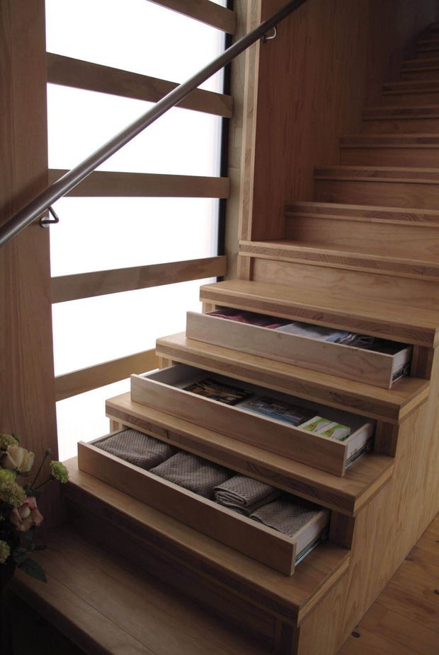 Interior view of a compact timber staircase with integrated pull-out drawers beneath several steps. The stairs and surrounding wall are clad in warm natural wood, with a simple metal handrail and tall frosted or overexposed glazing with horizontal timber framing alongside the stair.