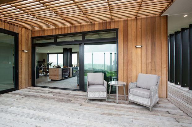 A covered outdoor terrace or deck with pale weathered timber decking, vertical timber wall cladding, and a slatted timber pergola overhead. Two light upholstered lounge chairs and a small round side table create a seating area beside large dark-framed glazed sliding doors. The glazing reveals a bright interior and distant landscape beyond. The space has a contemporary, warm, indoor-outdoor character.