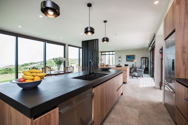 A spacious, open-plan contemporary kitchen with a large dark island, integrated sink, warm timber cabinetry, and built-in appliances. Black pendant lights hang above the island, while full-height glazing opens the room to a green rural landscape. A dining and living area is visible beyond the kitchen, with a vertical black slatted feature partition adding definition.
