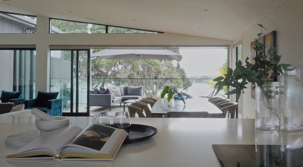 Open-plan contemporary living and dining area viewed from a kitchen island or benchtop. The bright interior features extensive floor-to-ceiling glazing, a large dining table with upholstered chairs, a grey outdoor-style lounge setting beyond, and a neutral kitchen surface in the foreground. Indoor plants and garden views add a relaxed connection to the outdoors.