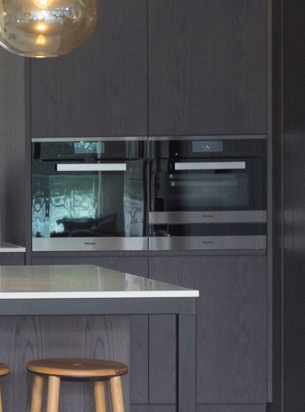 Close-up view of a contemporary kitchen with dark vertical-grain cabinetry, integrated stainless-steel ovens, and a pale stone island benchtop. A timber stool and glass pendant light add warm accents to the otherwise dark, refined scheme.