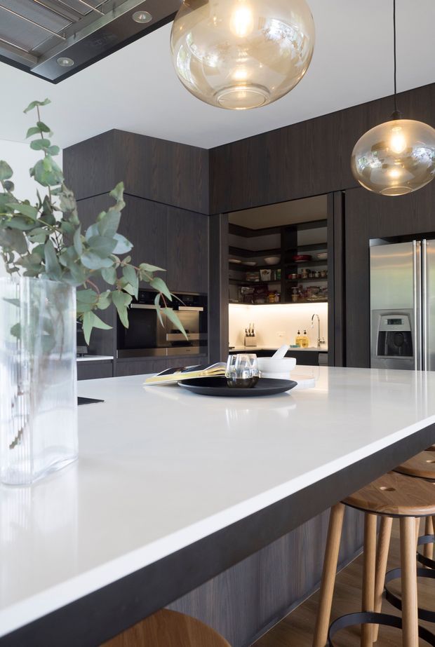 A sophisticated modern kitchen featuring dark timber cabinetry, integrated appliances, a large white island benchtop with seating, warm glass pendant lights, and softly illuminated open pantry shelving. Natural greenery and timber stools add warmth to the otherwise dark-and-white palette.