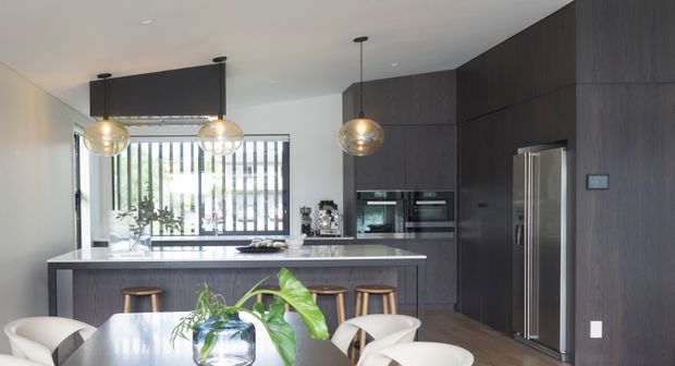 Contemporary open-plan kitchen and dining area with dark timber-look floor-to-ceiling cabinetry, a pale stone island benchtop, integrated appliances, pendant globe lights, and a dining table in the foreground. Large windows with vertical screening bring in abundant daylight.