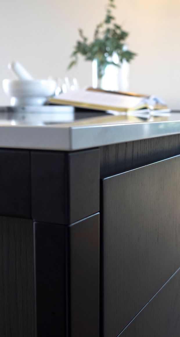 Close-up architectural interior detail showing dark, vertically grained built-in cabinetry with crisp geometric panel divisions and a thick pale stone benchtop. A softly blurred tabletop arrangement with a vase of greenery and serving objects sits above, creating a refined contemporary kitchen or dining joinery vignette.