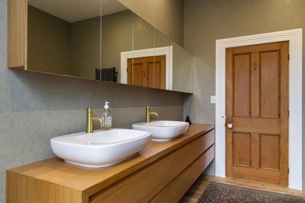 A compact, warm-toned bathroom with a long timber vanity featuring two white vessel basins, brushed gold wall-mounted taps, mirrored cabinetry above, and a small square-tile splashback. The room has textured taupe walls, white decorative door trim, and a traditional timber panel door. A patterned rug adds warmth near the doorway.