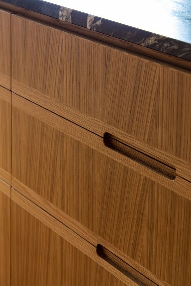 Close-up of richly grained mid-tone timber cabinetry with vertically fluted or reeded drawer fronts and integrated dark recessed pulls. A dark marbled stone benchtop is visible along the upper edge, highlighting refined custom joinery.