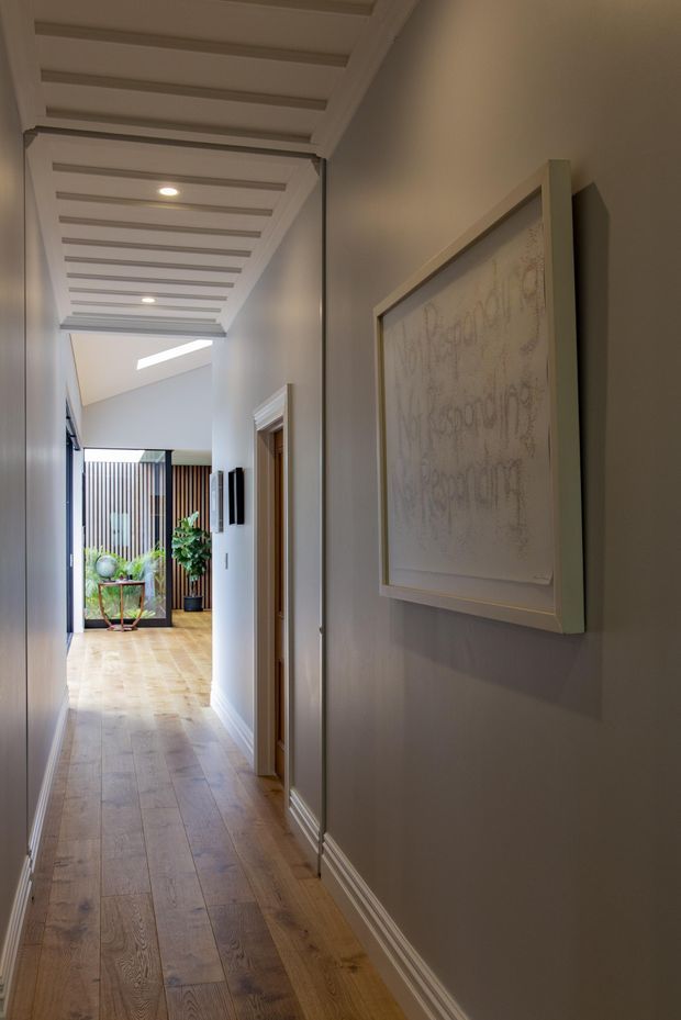 A long, refined residential hallway with warm timber flooring, pale painted walls, detailed white skirting boards, and a decorative coffered or battened ceiling with recessed downlights. A framed artwork hangs on the right wall, while the corridor leads toward a bright glazed entry or courtyard area with indoor plants and vertical screening.