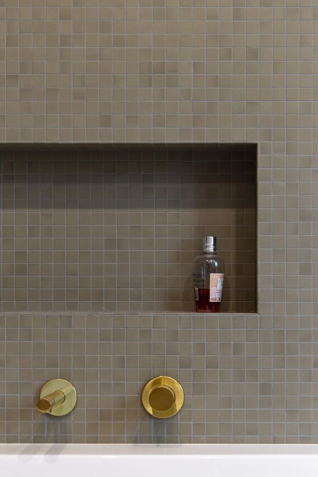 Close-up of a refined bathroom shower or bath wall clad entirely in small square taupe-grey tiles. A recessed tiled niche holds a soap dispenser, with two round brushed-gold controls below and the white edge of a bathtub visible at the bottom.