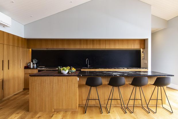 A warm, contemporary kitchen featuring extensive vertical timber cabinetry and island cladding, a long black stone benchtop, a dramatic black splashback, and four black bar stools. The open-plan space has pale walls, a white paneled ceiling, timber flooring, integrated appliances, and a simple linear layout.