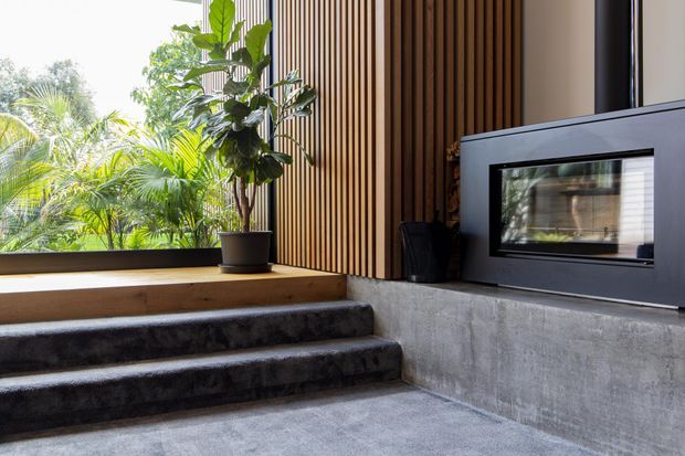 A contemporary indoor-outdoor transition space with broad concrete steps, a low raw concrete wall, vertical timber battens, lush tropical planting visible through glazing, and a built-in dark fireplace or firebox. The restrained material palette combines concrete, timber, and dark metal framing with strong natural light.