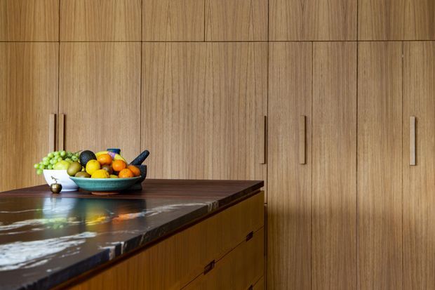 Close-up view of a refined kitchen with full-height vertical timber cabinetry, integrated tall storage, and a dark polished stone island benchtop. A fruit bowl sits on the island, adding a small colourful accent against the warm wood palette.
