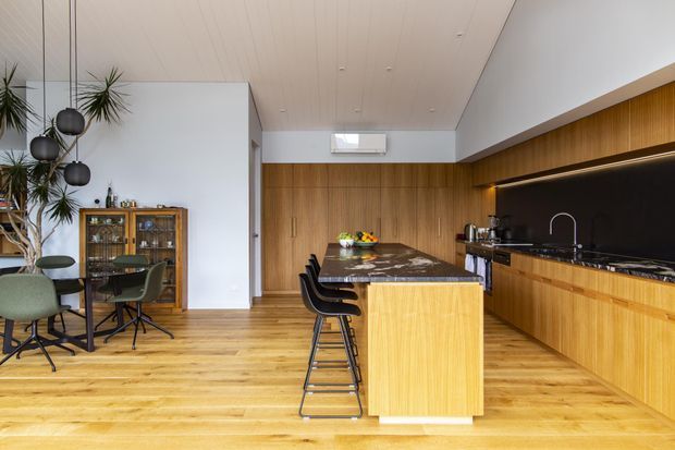 Open-plan contemporary kitchen and dining area with extensive warm timber cabinetry and flooring. A dark stone island with bar seating sits centrally, while the kitchen wall features integrated cabinetry, a dark splashback, under-cabinet lighting, sink and appliances. The adjoining dining area includes a glass table, upholstered chairs, pendant lighting and a timber display cabinet.