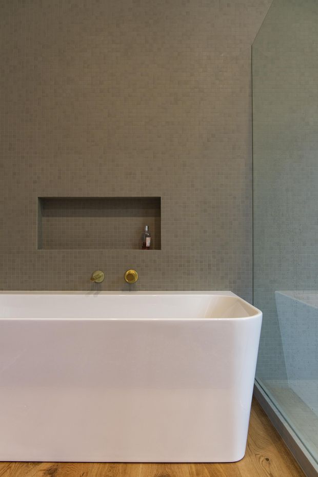 Contemporary bathroom featuring a freestanding white bathtub positioned beside a glazed shower enclosure. The walls are finished in small beige-grey mosaic tiles with a recessed wall niche, and brushed gold-toned bath fittings provide a subtle accent. Warm timber flooring adds contrast.