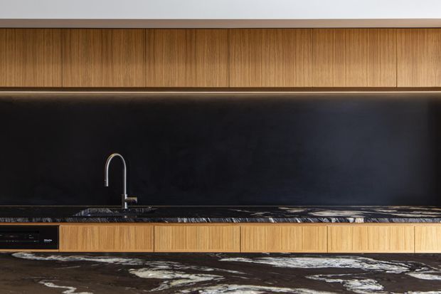 A sleek contemporary kitchen wall with warm vertical-grain timber cabinetry, a continuous matte black splashback, integrated under-cabinet lighting, a minimalist curved faucet, and a dramatic dark stone benchtop with pale veining. The composition focuses on the fixed kitchen joinery and finishes.