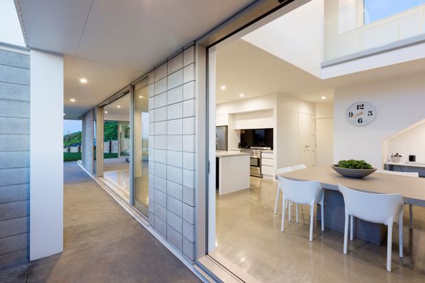 A bright contemporary open-plan interior viewed from a covered outdoor walkway, with a white kitchen, dining island, polished concrete flooring, extensive sliding glass doors, and a double-height living area with an upper-level opening. The exterior walkway and garden are visible through the glazing.