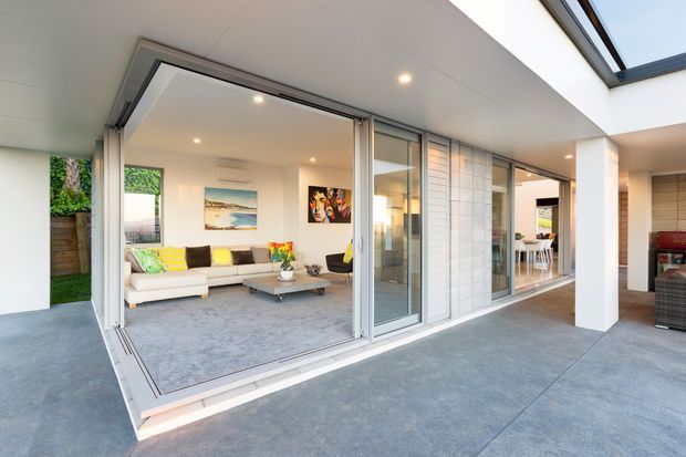 Bright contemporary home interior opening onto a covered outdoor entertaining area. Large aluminium-framed sliding glass doors connect a carpeted living room with a broad concrete-paved terrace. The living room contains a light sofa, low coffee table, colourful cushions and wall art, while a dining area and outdoor barbecue zone are visible beyond. White walls, recessed lighting and a crisp open-plan layout create an airy, modern feel.