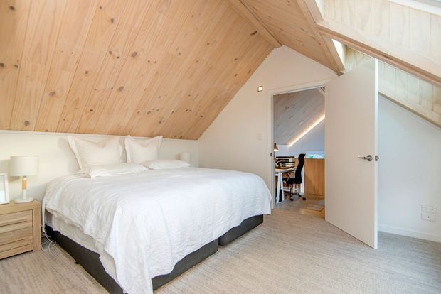 Bright attic-style bedroom with a low-sloped, exposed timber-lined ceiling, white walls, a large white bed, pale carpet, and simple light-wood bedside furniture. An open door reveals a small adjacent workspace with timber finishes and glazing. The room has a clean, airy, minimalist character.