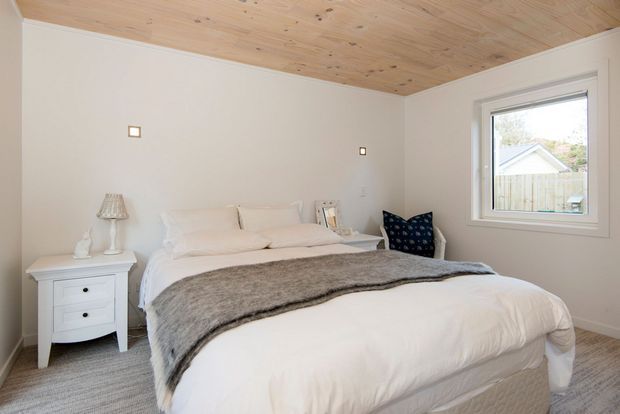 A calm, compact bedroom with a neatly made bed, white bedding, a taupe-grey throw, white bedside furniture, and a navy patterned accent chair. The room has warm light-wood ceiling boards, pale neutral walls, carpeted flooring, simple square wall lights, and a white-framed window.