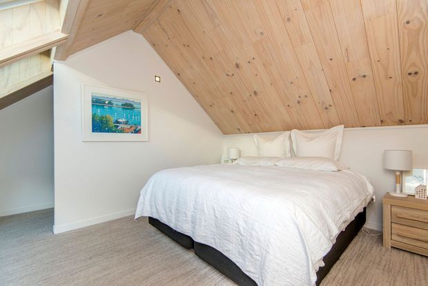 A bright attic bedroom with a sloped, natural timber-lined ceiling, white walls, a neatly made bed, light carpet, and a simple timber bedside table. A framed artwork and small bedside lamps add understated detail. The low roofline and exposed wood ceiling create a warm, compact retreat.