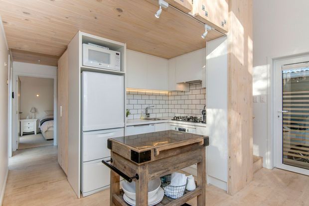 Bright compact kitchen with white cabinetry, a white subway-tile splashback, integrated refrigerator and microwave, gas cooktop, and a rustic timber island with a dark stone benchtop. The kitchen is within an open-plan timber-lined interior, with a bedroom visible through the hallway opening and a glazed exterior door at right.
