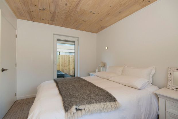 A bright, compact bedroom with a white upholstered bed dressed in white bedding and layered with a grey textured throw. The room has pale walls, a warm natural timber plank ceiling, simple white bedside furniture, and a narrow window looking toward a fenced exterior area. The overall design is calm, light, and understated.